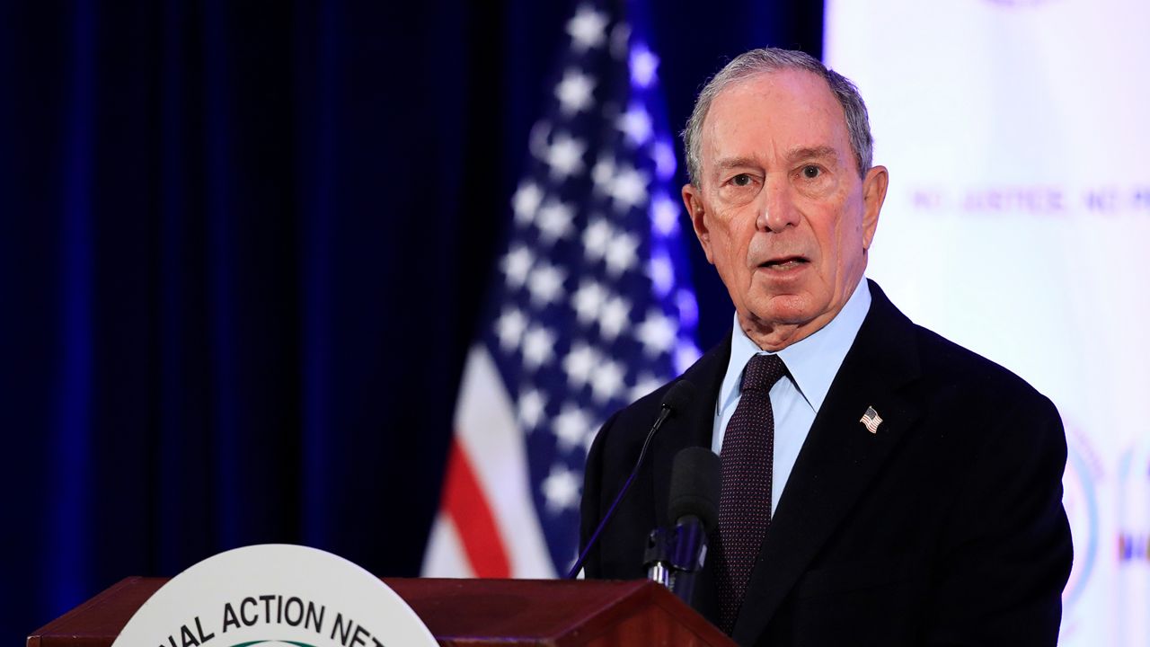 Michael Bloomberg, wearing a black suit jacket, a white dress shirt, and a dark purple tie, stands behind a black microphone attached to a wooden brown podium, upon which a white circular sign is affixed. A navy blue curtain and a red-white-and-blue American flag stand in the background.