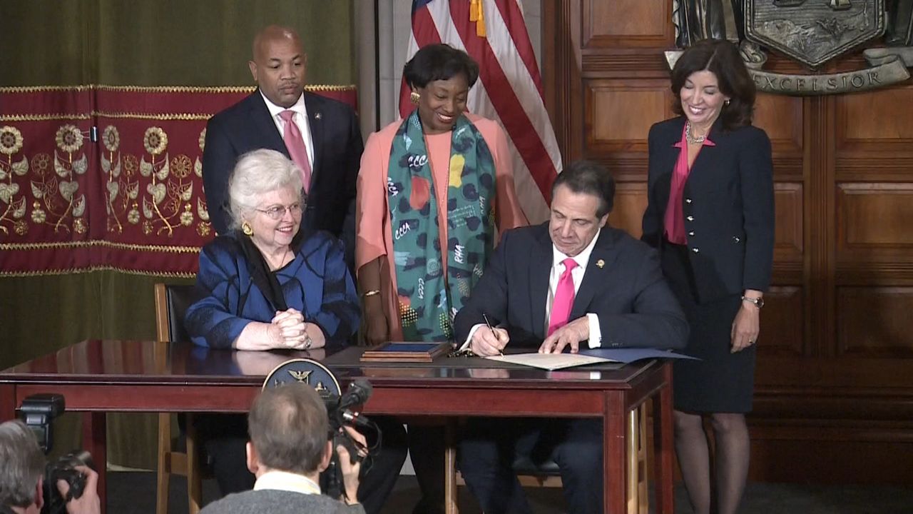New York Gov. Andrew Cuomo, right foreground, wearing a black suit jacket, a white dress shirt, and a pink tie, sits behind a wooden brown desk. He holds a black pen near a white sheet of paper. People wearing suit jackets and dresses stand behind him.