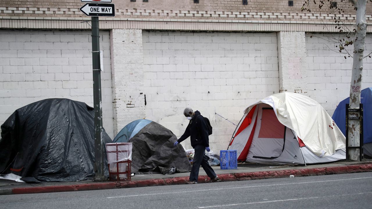 In this March 20, 2020 photo a man covers his face with a mask as he walks past tents on skid row, in Los Angeles. (AP Photo/Marcio Jose Sanchez)