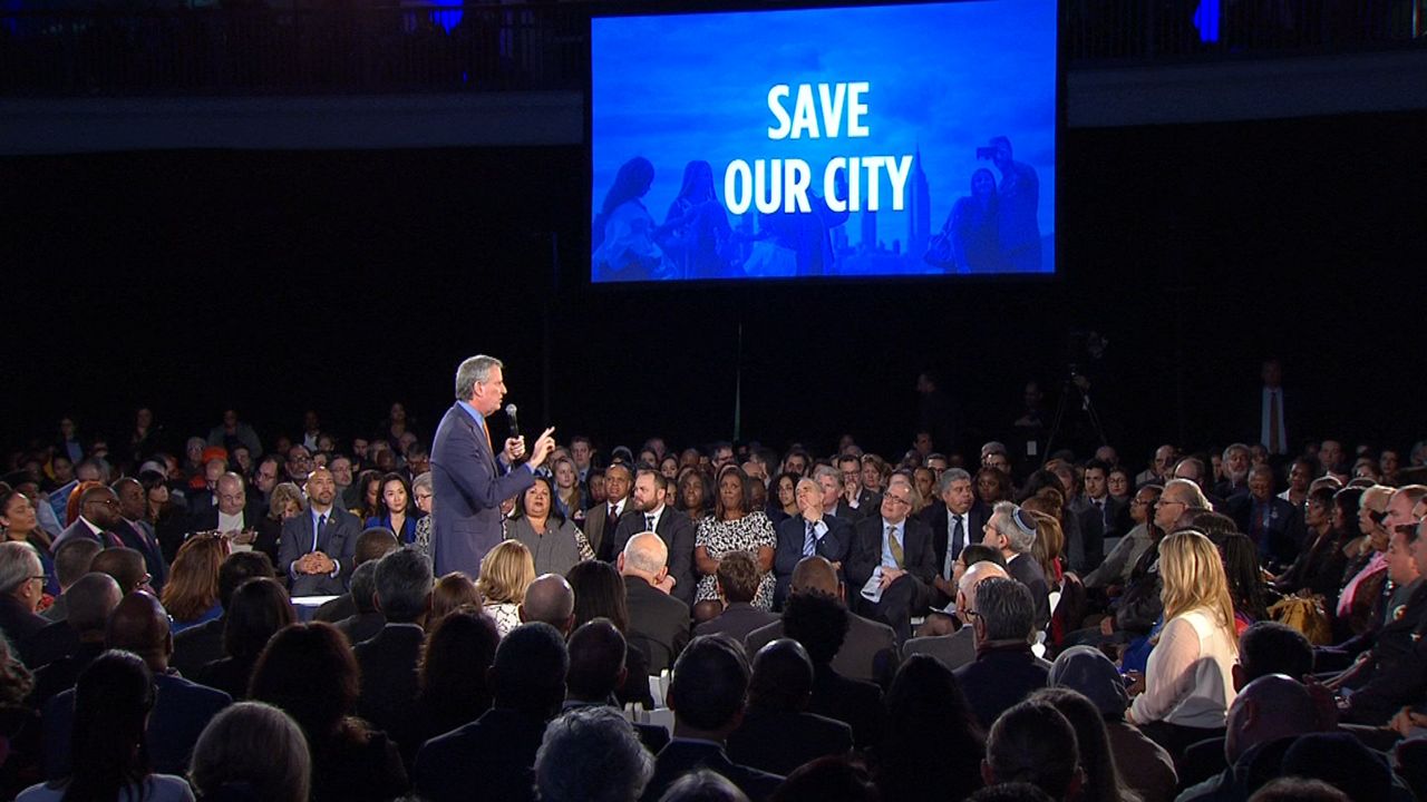 Mayor Bill de Blasio, left, foreground, wearing a navy blue suit jacket, stands amid a crowd of people during his State of the City address. A giant blue screen in the stance features white text that reads "Save Our City."