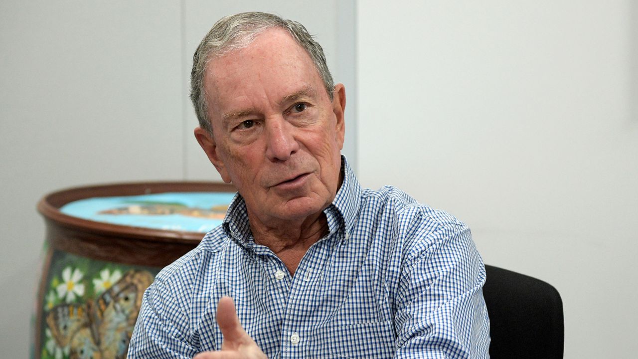 Michael Bloomberg, wearing a plaid dress shirt, sits in a black chair near a white wall.