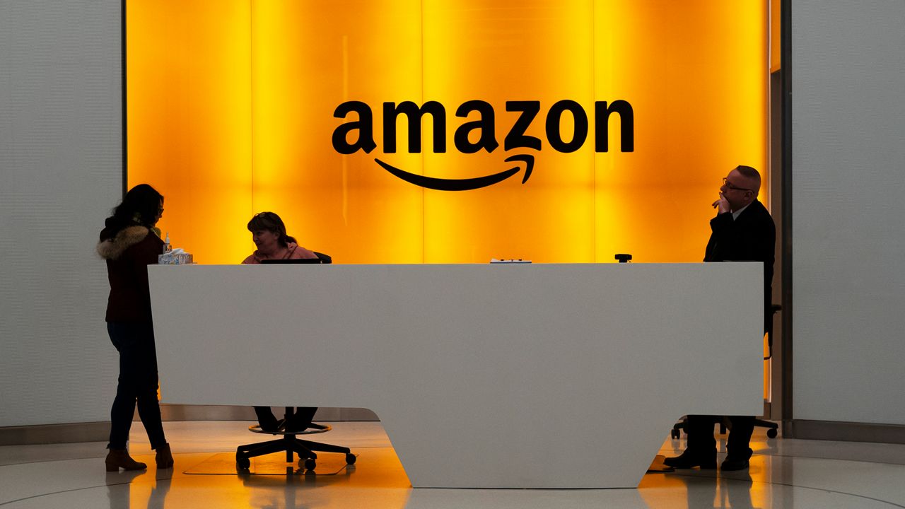 Black letters spell out "Amazon" against an orange-lit screen in an office. Three people stand and sit behind a white desk.