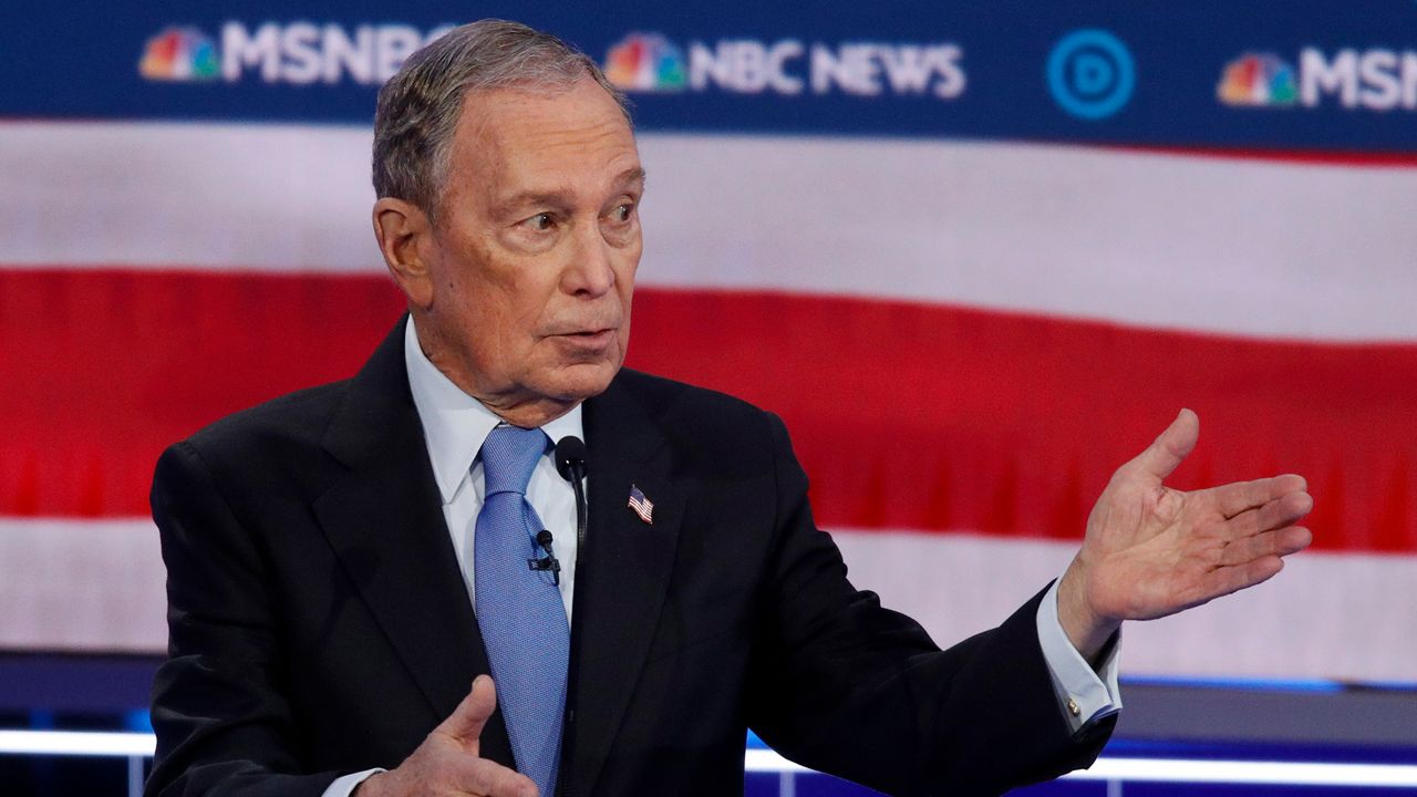 Michael Bloomberg, wearing a black suit jacket, a white dress shirt, and a sky blue tie, stands behind a background of an American flag and MSNBC and NBC News logos at the Democratic presidential debate in Las Vegas, Nevada.