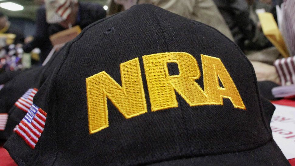 A black cap with an American flag printed on its right side and the NRA logo in bold yellow text on the front of the cap.