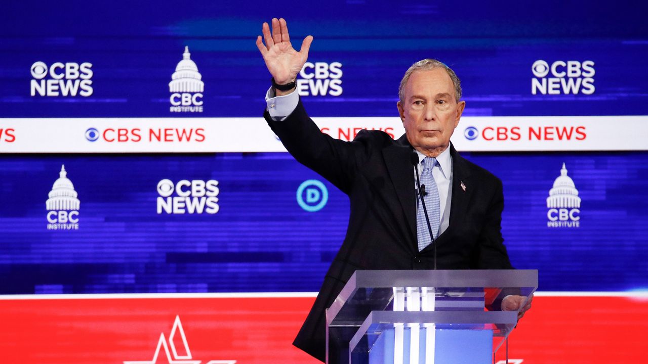 Michael Bloomberg, wearing a black suit jacket, a white dress shirt, and a sky blue tie, stands behind a clear glass podium in front of a blue, white, and red banner at a Democratic presidential primary debate at the Gaillard Center in Charleston, South Carolina.
