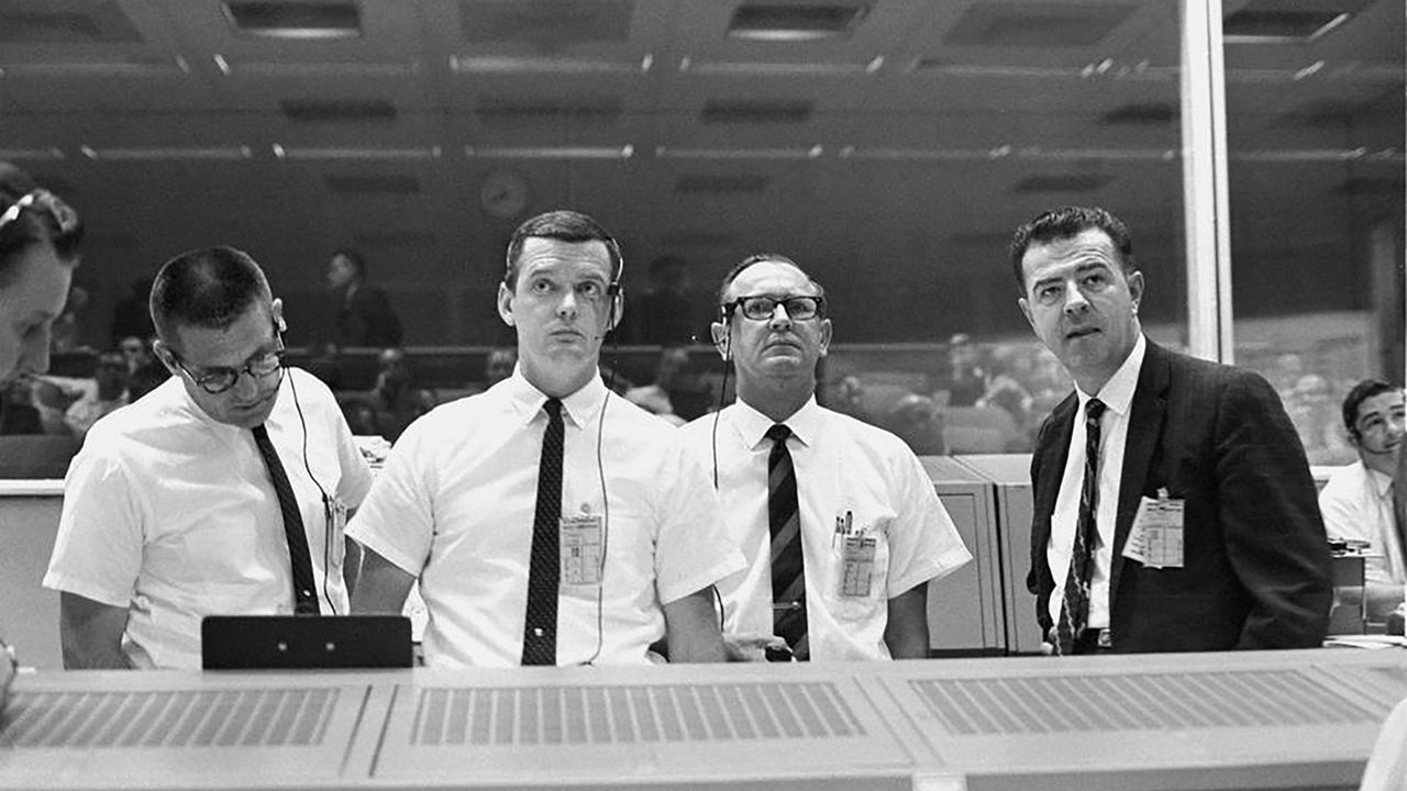 tanding at the flight director's console, viewing the Gemini-10 flight display in the Mission Control Center on July 18, 1966, are (left to right) William C. Schneider, Mission Director; Glynn Lunney, Prime Flight Director; Christopher C. Kraft Jr., MSC Director of Flight Operations; and Charles W. Mathews, Manager, Gemini Program Office. (NASA)