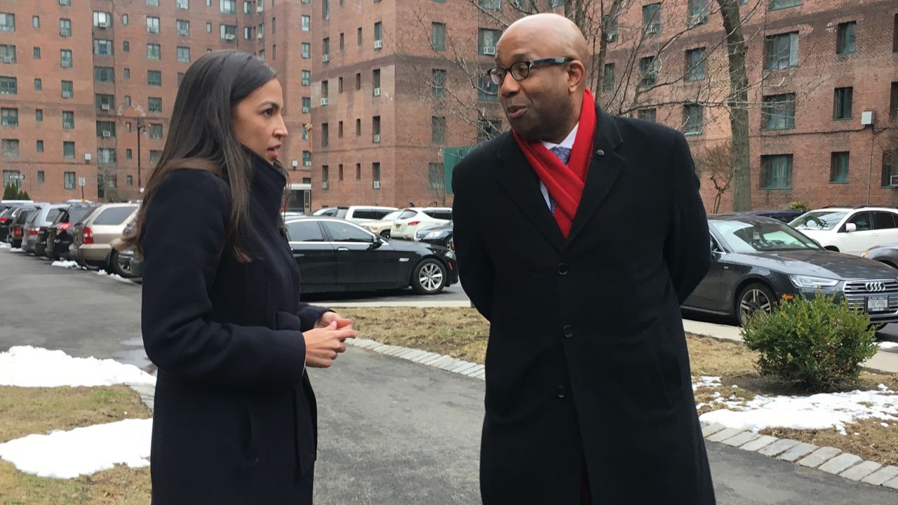 Rep. Alexandria Ocasio-Cortez, left, wearing a black coat. NY1's Errol Louis, right, wearing a black coat. They walk in a crosswalk. A black SUV is parked about five feet to the right of Ocasio-Cortez, in front of a bed of snow, about two feet tall and two feet wide.