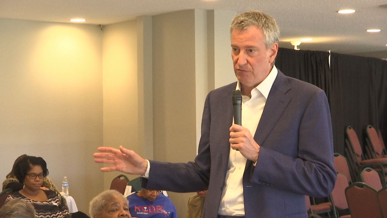 New York City Mayor Bill de Blasio wearing a white dress shirt and a navy blue blazer while holding a black microphone in a hall.