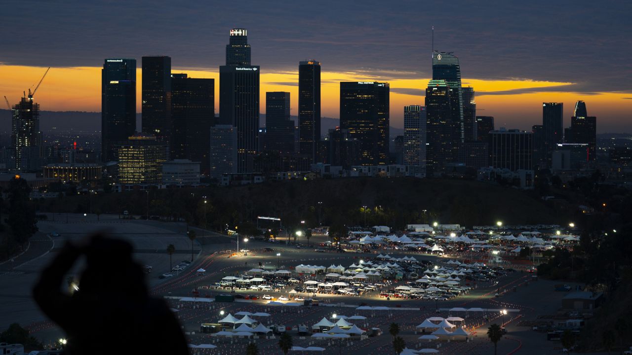 A spectator stands on a hilltop overlooking downtown Los Angeles as motorists line up for COVID-19 vaccinations and testing in the parking lot of Dodger Stadium in Los Angeles on  Jan. 27, 2021. (AP Photo/Jae C. Hong)