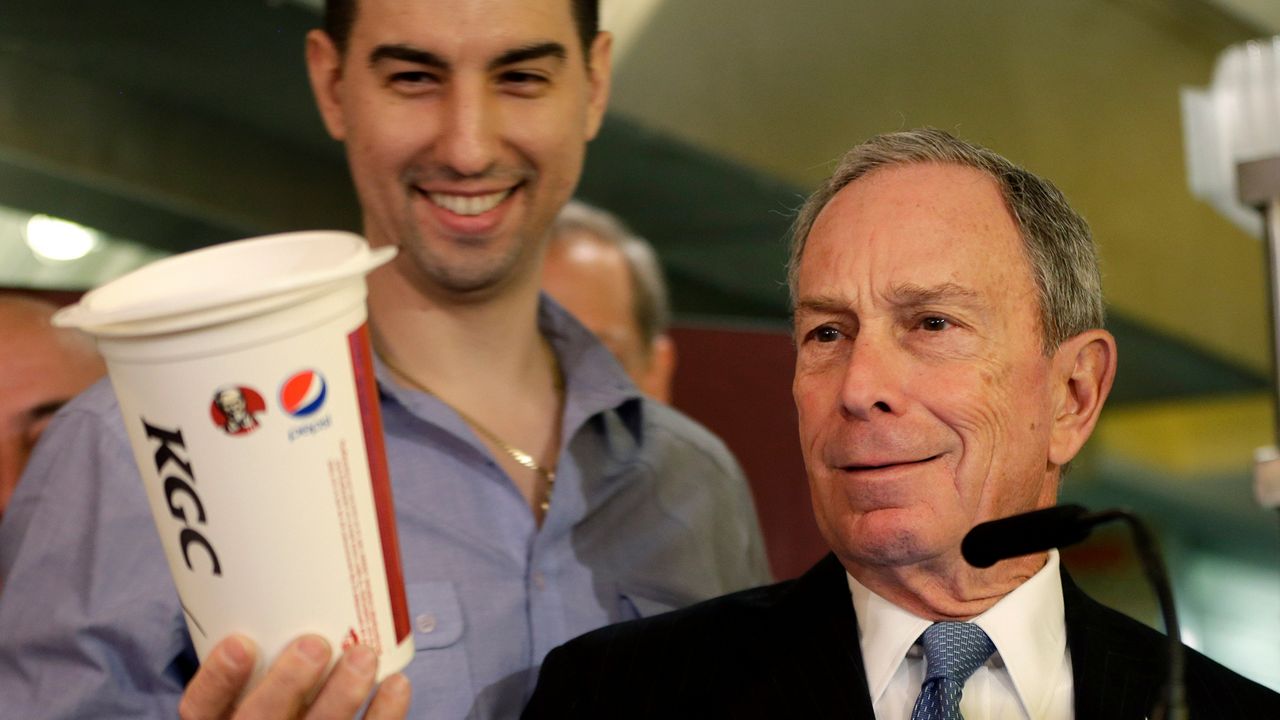 Mayor Michael Bloomberg, wearing a black suit jacket, a white dress shirt, and a blue tie, stands behind a black microphone and a dark lectern while holding up a white KFC up in his right hand. A man, wearing a blue dress shirt, stands behind him.