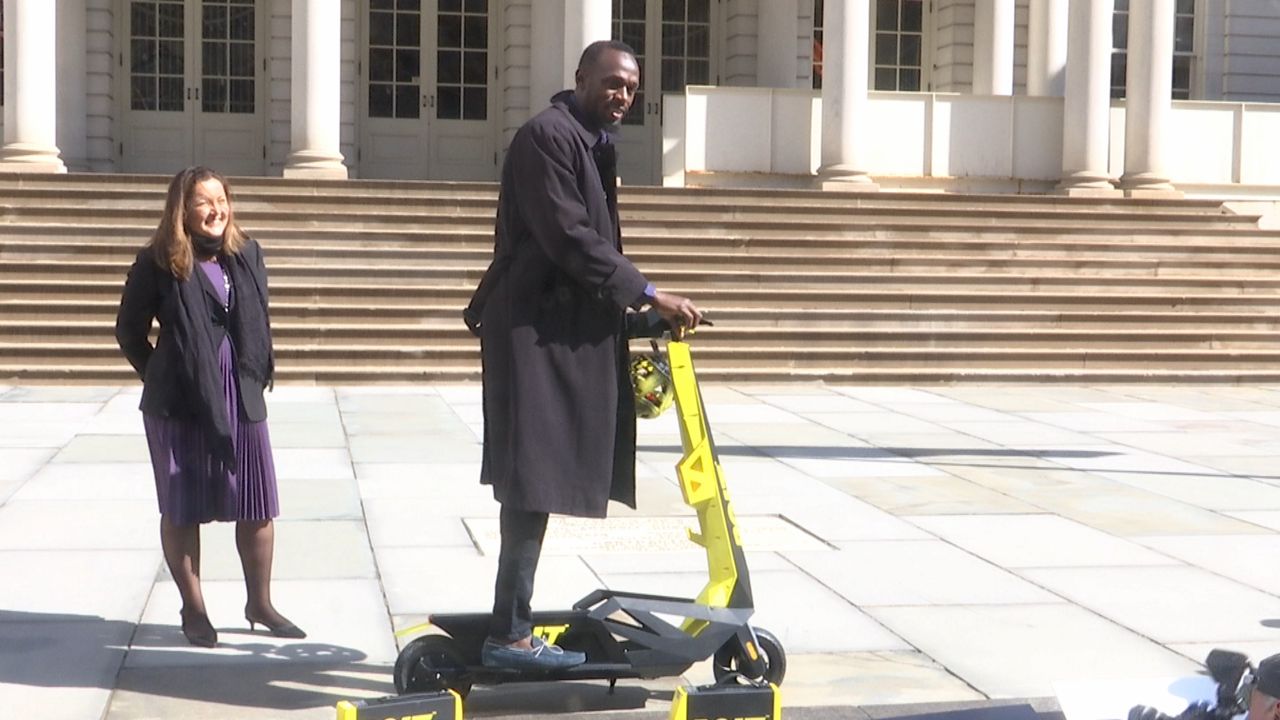Usain Bolt, wearing a black coat, black pants, and black sneakers, stands on an e-scooter on white steps. A person wearing a black coat and a purple dress stands behind him.