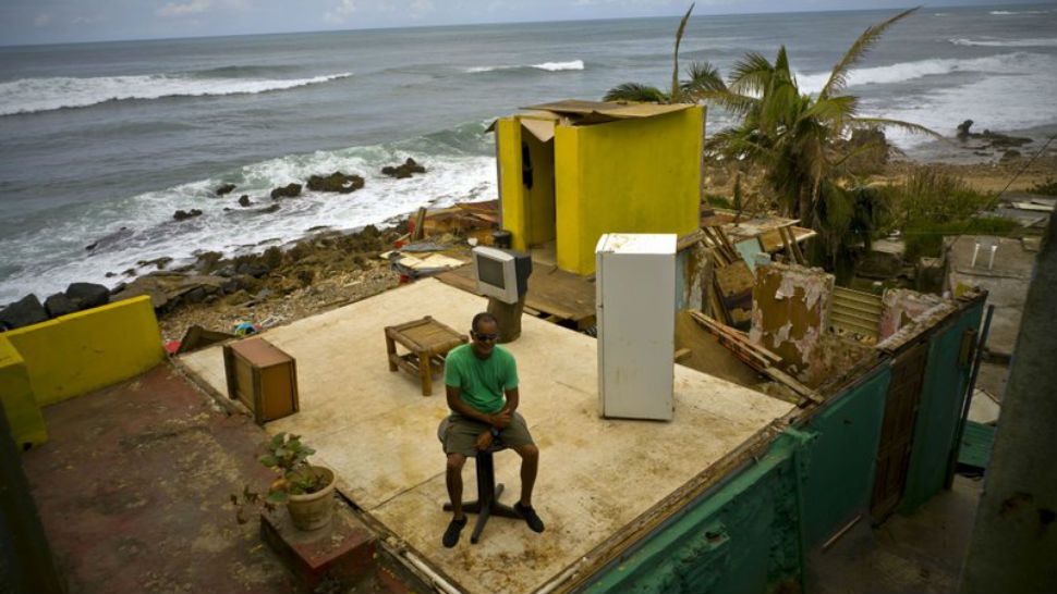 FILE - In this Oct. 5, 2017 file photo, Roberto Figueroa Caballero sits on a small table in his home destroyed by Hurricane Maria, in La Perla neighborhood on the coast of San Juan, Puerto Rico. Maria destroyed up to 75,000 homes and damaged 300,000 more, causing an estimated $31 billion in damage to housing alone, said the island’s housing secretary, Fernando Gil.(AP Photo/Ramon Espinosa, File)
