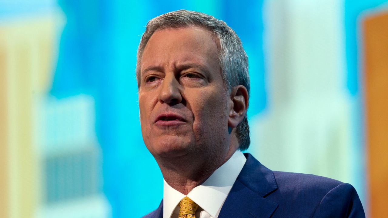 New York City Mayor Bill de Blasio, wearing a navy blue suit jacket, a white dress shirt, and a yellow tie.