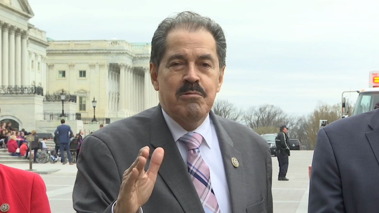 A man, wearing a grey suit jacket, a sky blue dress shirt, and a pink tie, stands near Capitol Hill.