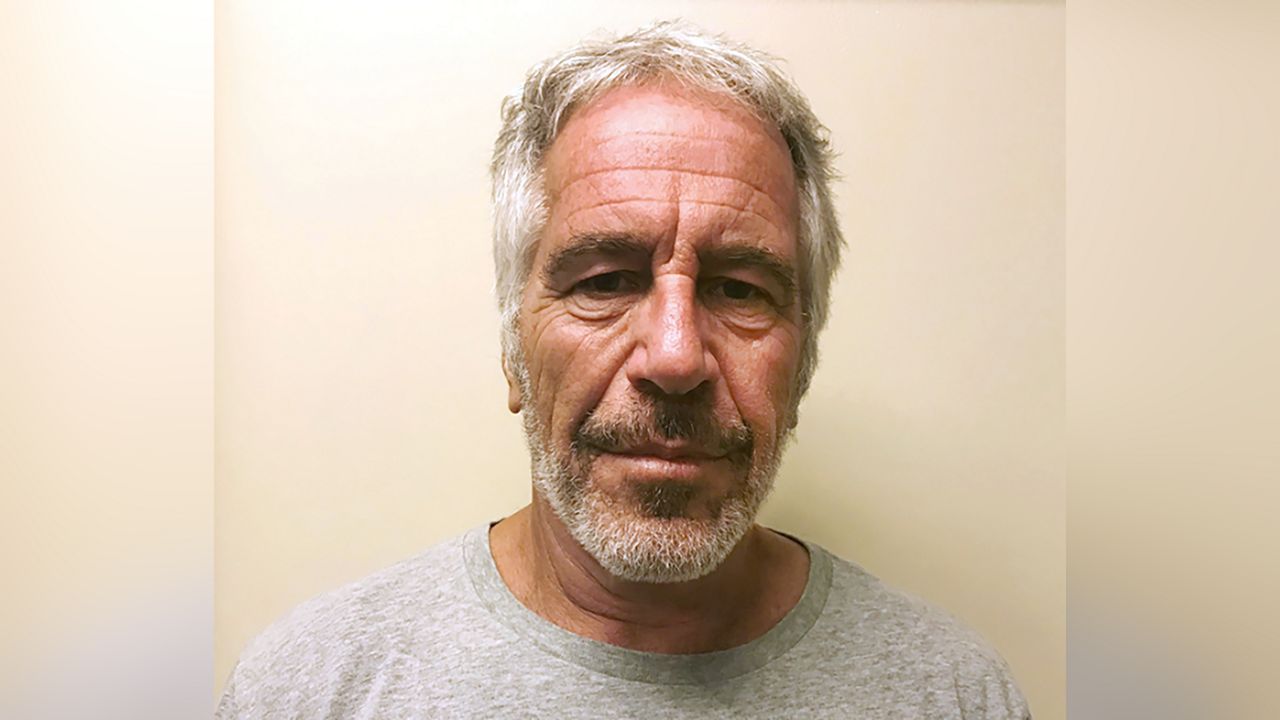 Jeffrey Epstein, wearing a white T-shirt, standing against a beige wall.