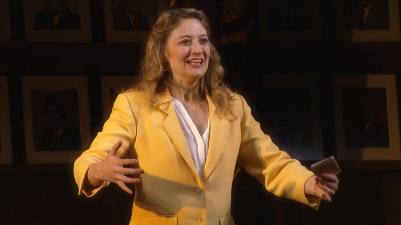 A woman, wearing a white dress shirt and a yellow blazer, stands in front of a set of black-and-white photo frames and two American flags.