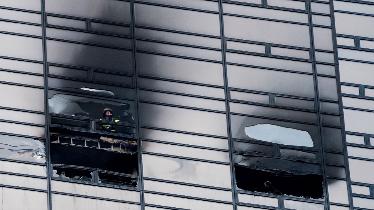 A building with scorch marks near two windows. A firefighter in a black and green uniform looks out from a window.