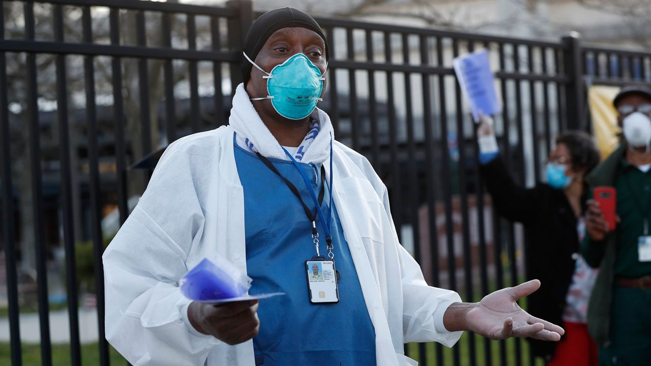 A Brooklyn VA hospital employee, wearing a white doctor's coat, blue medical scrubs, and a light blue mask, stands outside the hospital.