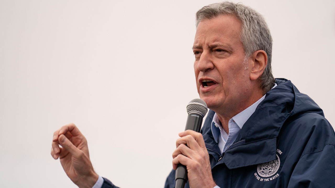 An April 9, 2021, file photo of Mayor Bill de Blasio, wearing a navy blue windbreaker and holding a black microphone, speaking before the ribbon cutting and seasonal opening of the Coney Island amusement park area in Brooklyn