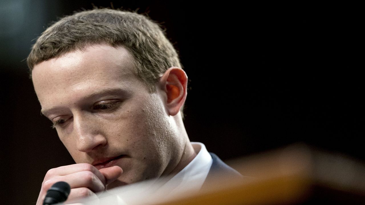 Mark Zuckerberg, wearing a suit and a dress shirt, holds his right hand near his bottom lip. A thin black microphone is inches away from his hand.