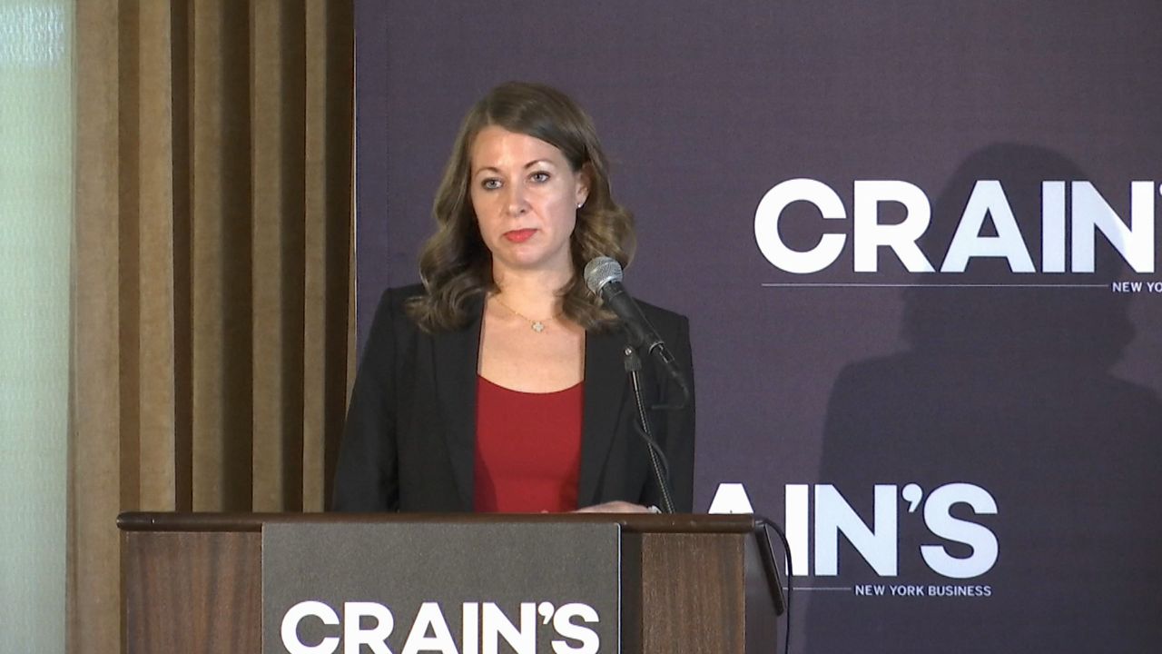 A woman, wearing a red shirt and a black blazer, stands a brown lectern. She speaks into a black microphone. A black sign, with white text that reads "Crain's," is placed on the center of the lectern. Black wallpaper with the same sign is behind the woman.