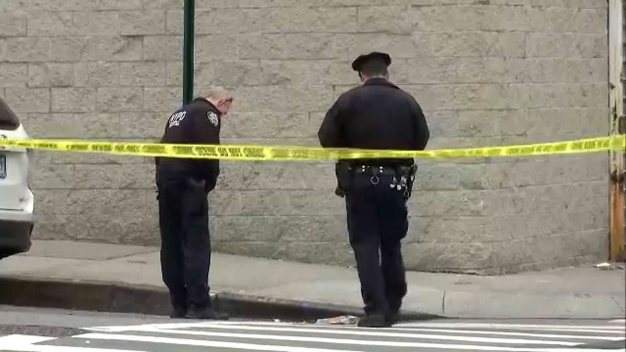 Two people in black NYPD uniforms stand near a curb, about two feet away from a grey brick wall. Yellow police tape is about teen feet behind them.