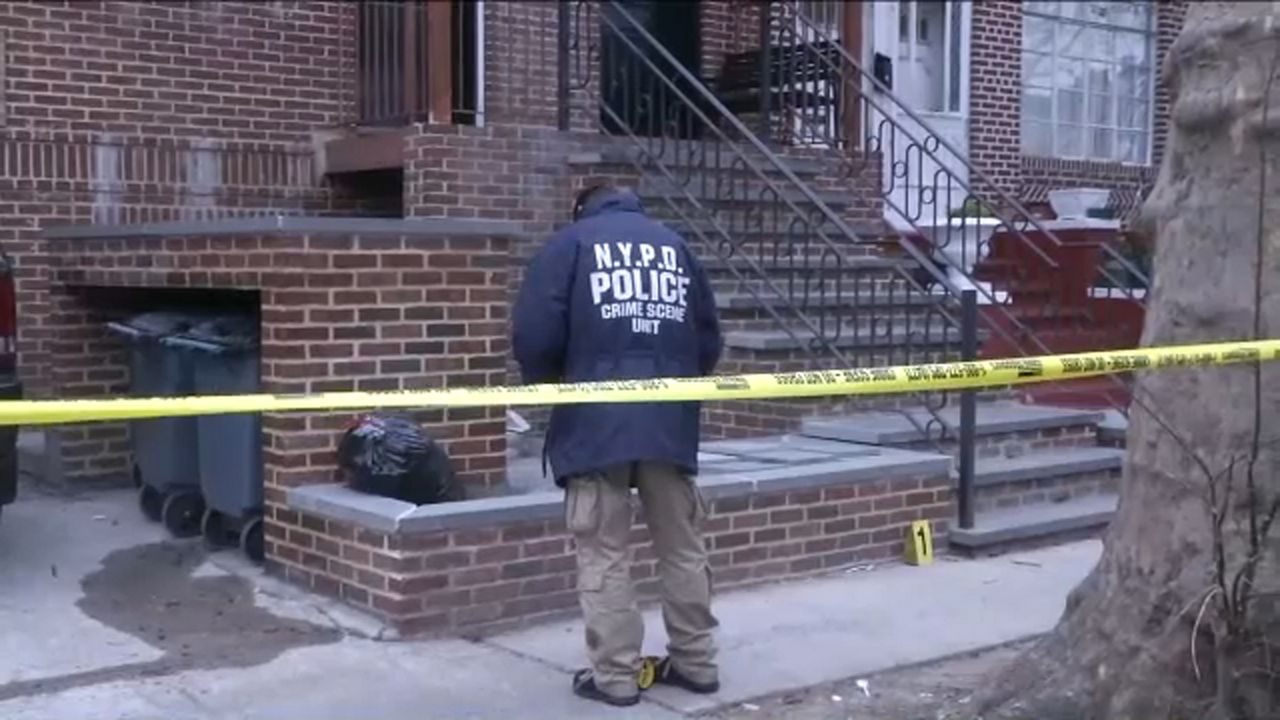 A person in a blue NYPD jacket and brown khaki pants near a red-brick staircase. Yellow police tape is about four feet behind him.