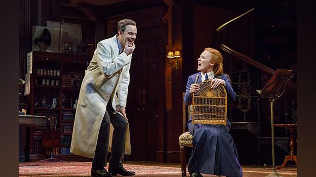 A man, left, in a white coat, wearing black pants and black dress shoes, holds his jaw while kneeling about two feet away from a woman in a blue dress, right, who holds a bookcase.