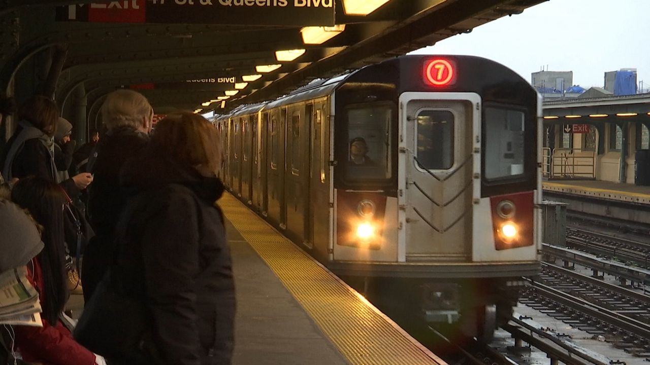 A silver 7 train, right, pulls into a station. A crowd of people wearing coats stand near the platform.