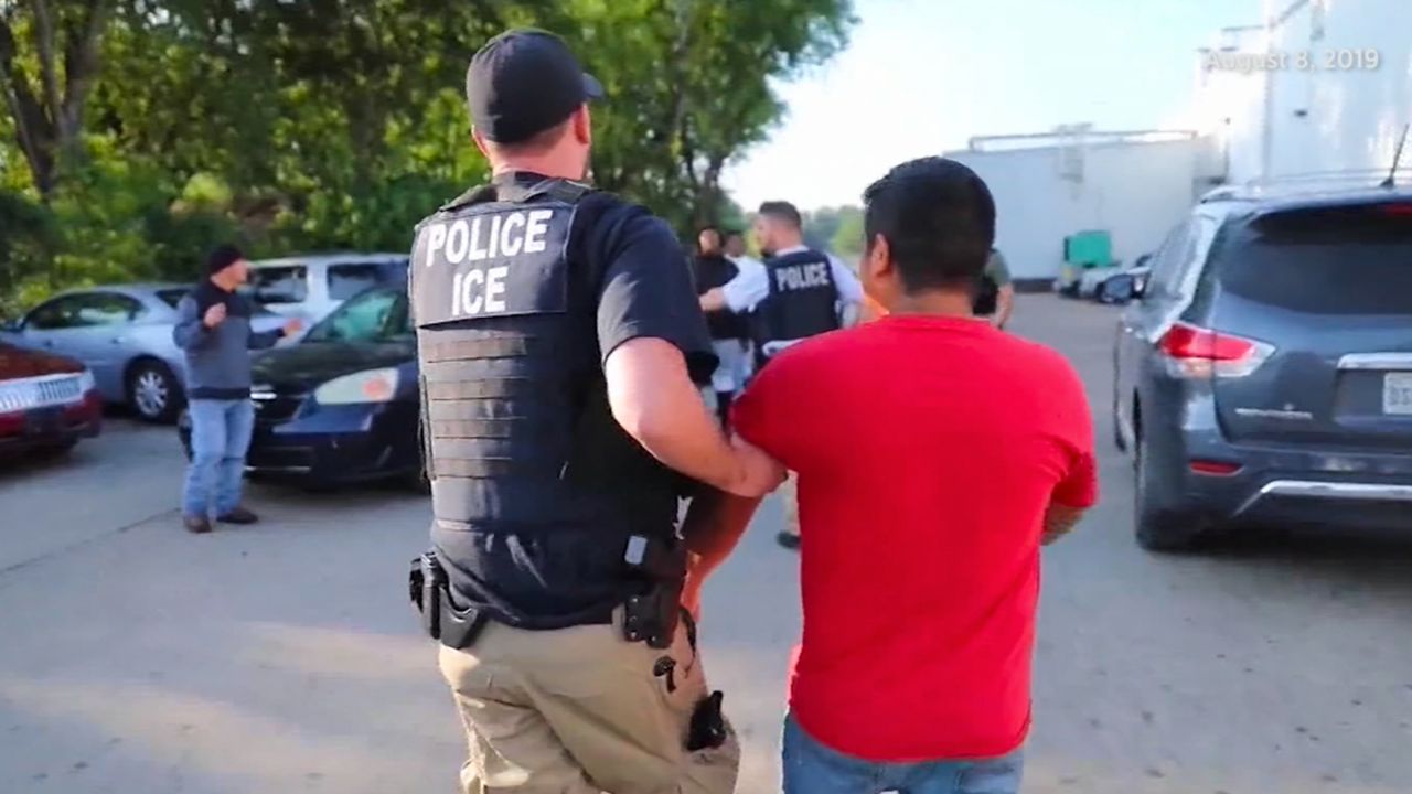 An agent for Immigrations and Customs Enforcement is shown from the back, wearing a bulletproof vest that says "POLICE ICE," escorting a man in a red shirt to a law enforcement vehicle.