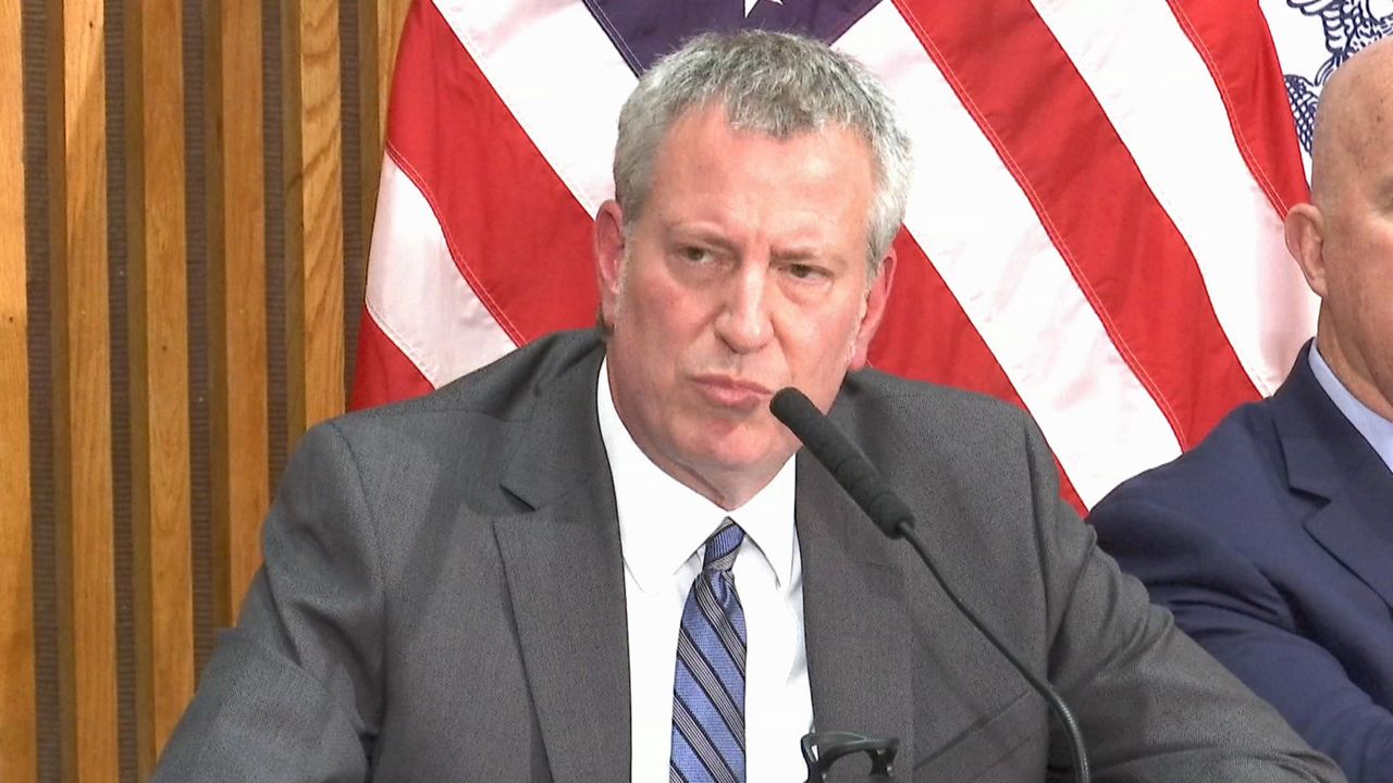 New York City Mayor Bill de Blasio, wearing a white dress shirt, a grey suit jacket, and a blue-and-grey-striped tie, speaks into a thin black microphone while sitting in front of an American flag.