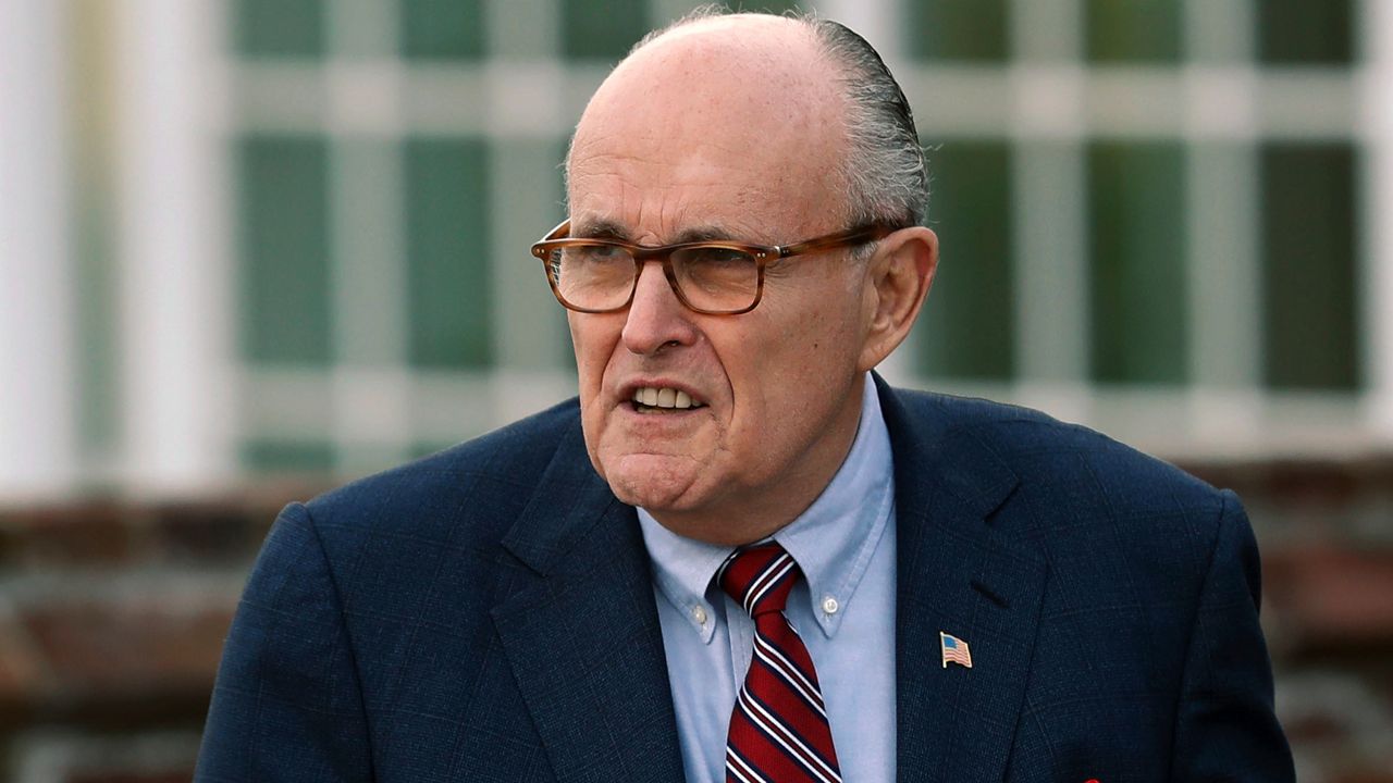 Rudy Giuliani wearing square glasses, a black suit jacket, a sky blue dress shirt, and a red tie with stripes.