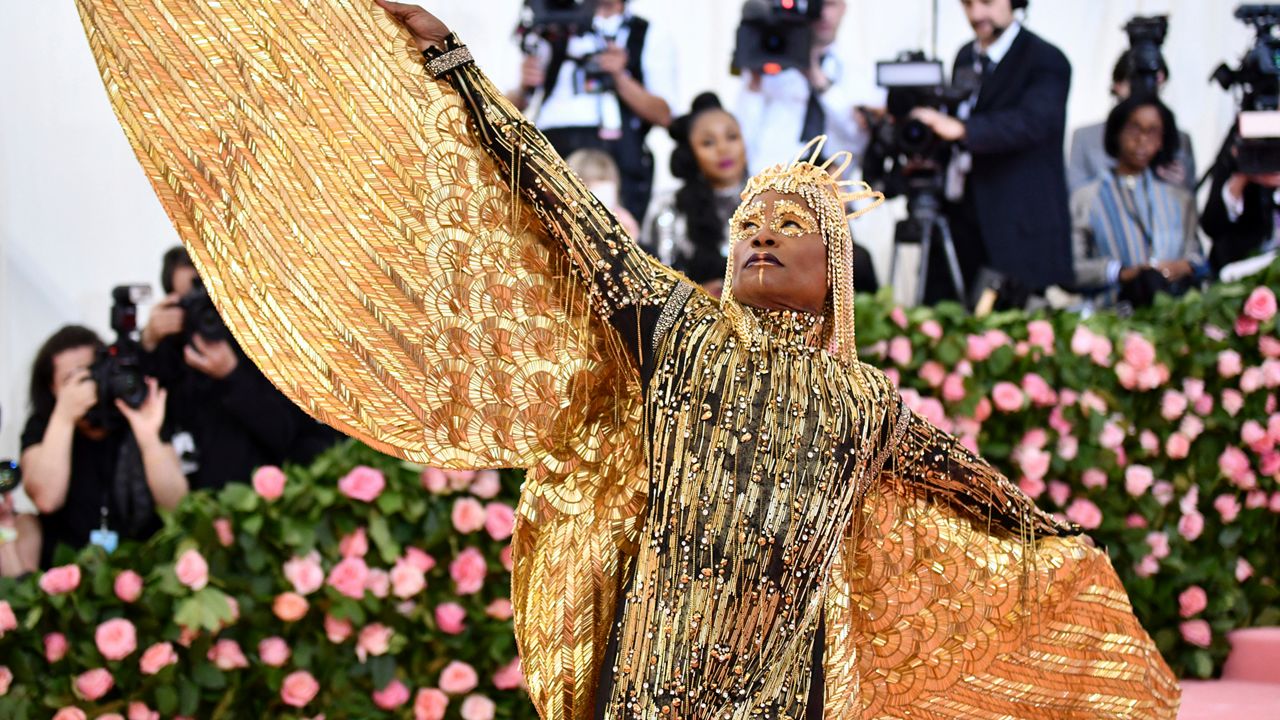 Billy Porter, wearing a gold crown and dress with an outstretched gold wing.