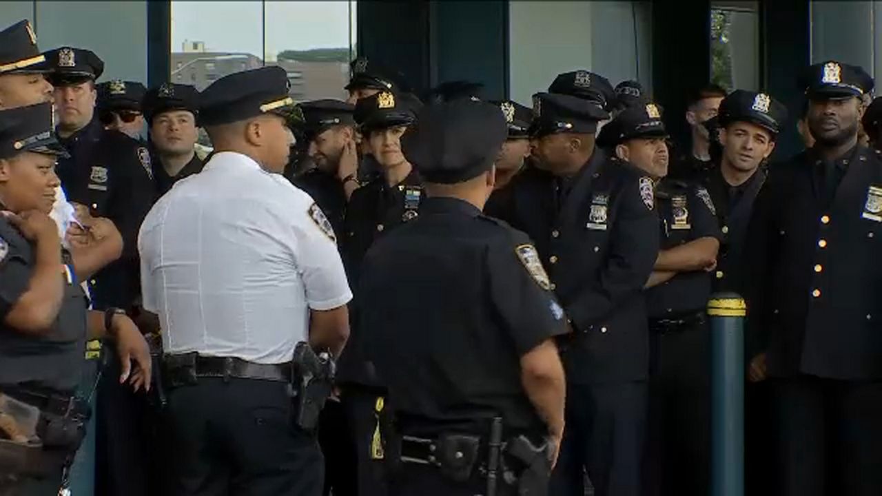 Men in women in black NYPD uniforms. A man in a white NYPD shirt is second-from-the-left.