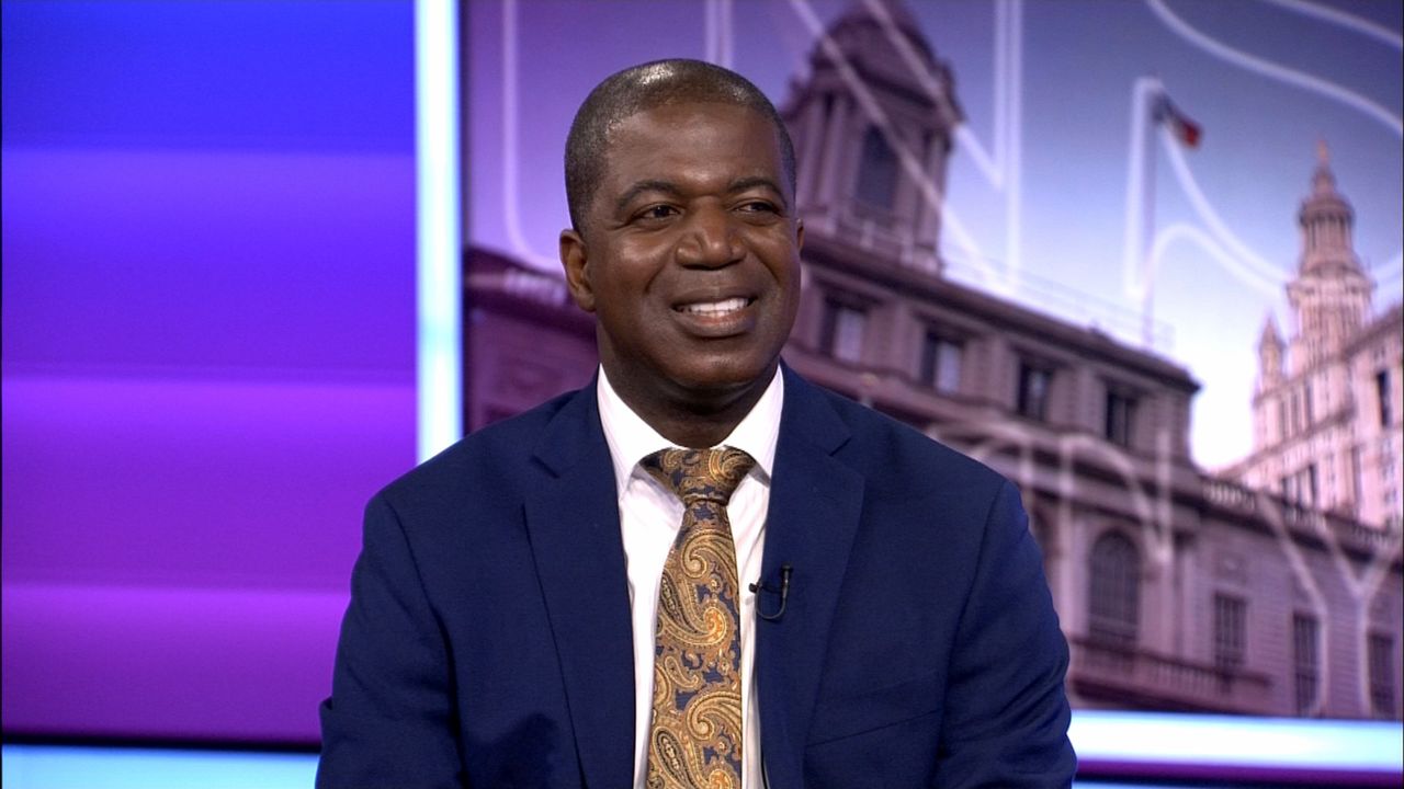 A man, wearing a navy blue blazer, a white dress shirt, and a bronze tie, sits against a purple background on the set of NY1 political show 