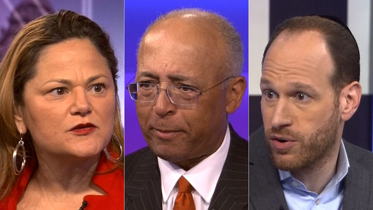 From left to right on the set of NY1 political show Inside City Hall: A woman, wearing a red blouse and gold earrings; a man, wearing square glasses, a black suit jacket, a white dress shirt, and a red tie; a man, wearing a sky blue dress shirt and a black blazer.