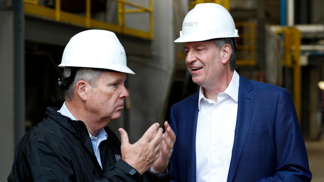New York City Mayor Bill de Blasio, right, wearing a blue blazer, a white dress shirt, and a white hard hat, stands a few inches away from a man, left, wearing a black coat, a white hard hat, and a sky blue dress shirt. Metal railguards for high platforms stand behind them.