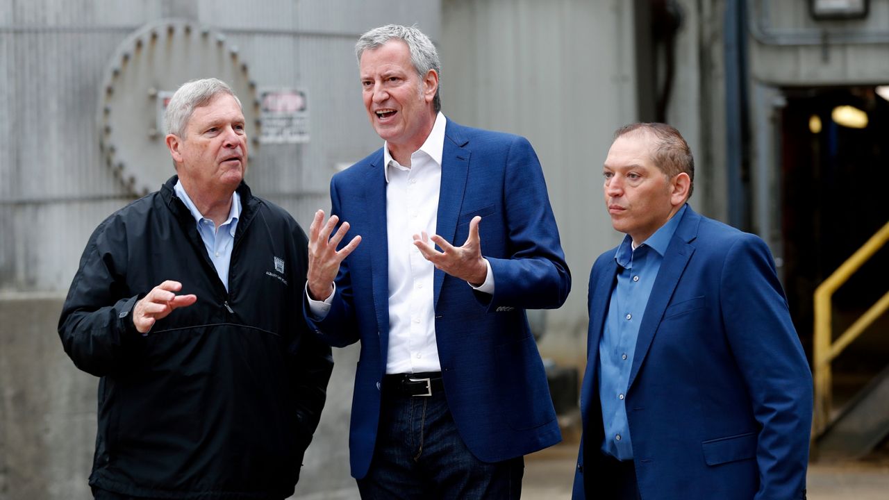 New York City Mayor Bill de Blasio, center, wearing a navy blue blazer, a white dress shirt, and black jean pants, stands next to two people.