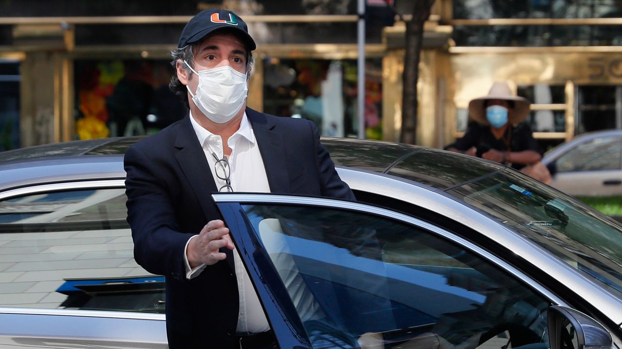 Michael Cohen, wearing a black hat, a white face mask, a black blazer, and a white dress shirt, upon which square, black-framed glasses hang, stands in between the passenger seat of a car and the door in Manhattan.