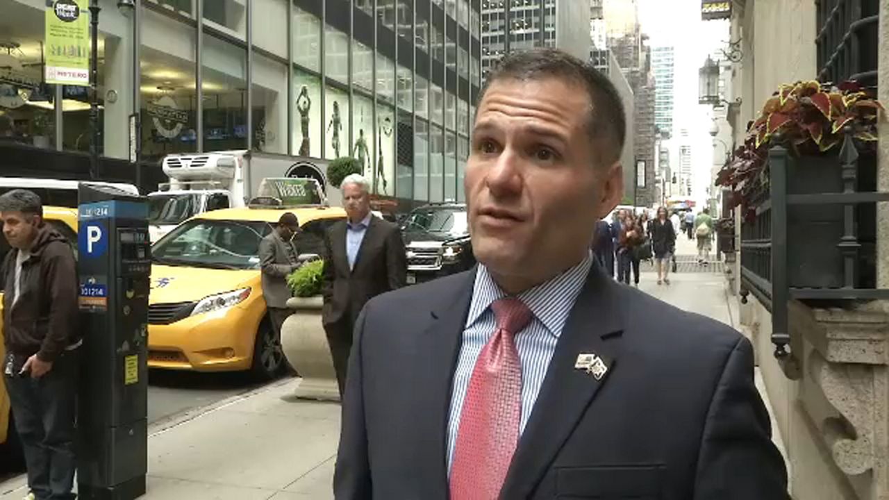 A man wearing a black suit jacket, a plaid dress shirt, and a pink tie stands on a city sidewalk. A line of parked cars are to his right.
