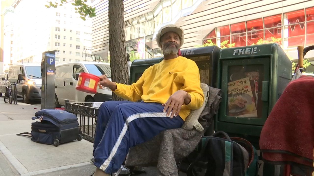 A man, wearing a yellow sweatshirt, a hat, and blue sweat pants with a white stripe, hold a can while sitting on a grey coat up against three green newspaper cans.