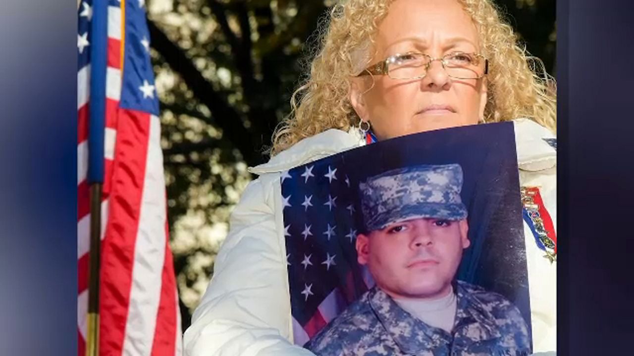A woman wearing glasses and a white jacket holds a photo while standing in front of an American flag.