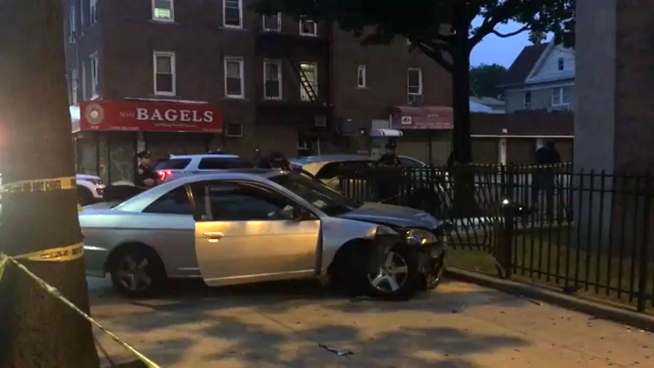 A silver car, with a door open, near a black pole that is laid out on a sidewalk in front of a black gate.