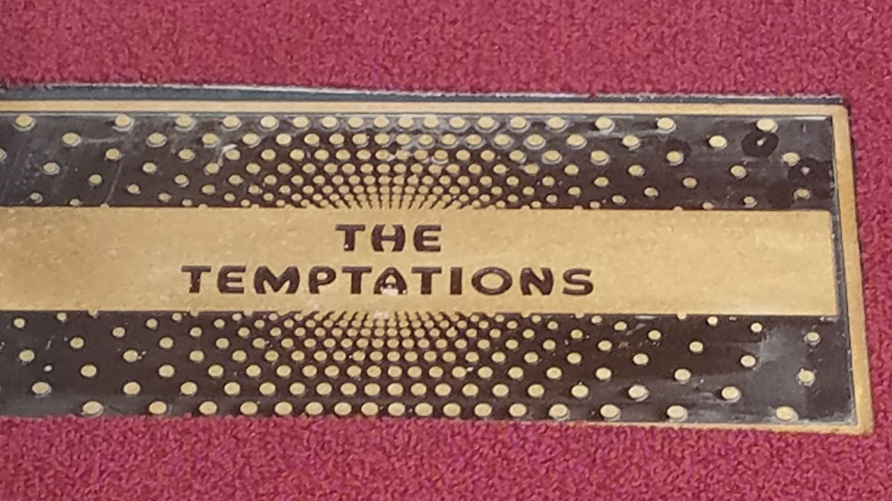 "The Temptations" written in black text on a gold slab on a black-and-gold plaque resting on red carpet. A person's white-and-black sneaks sits on the plaque.