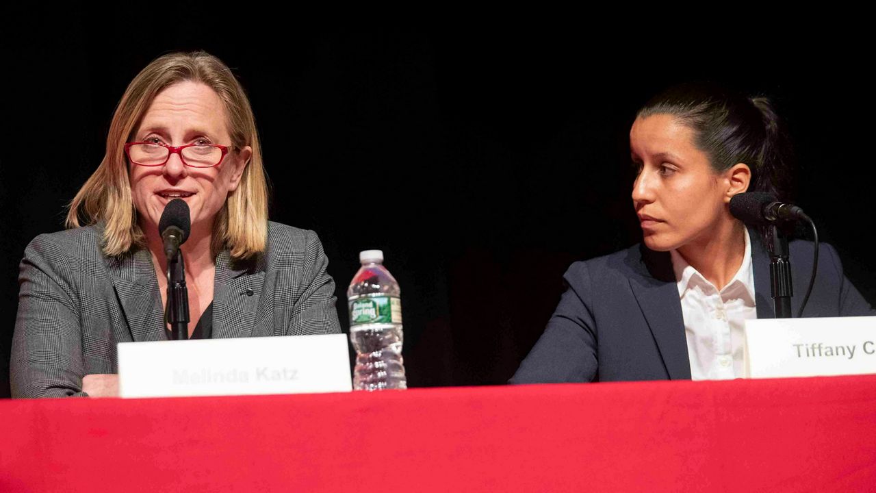 A woman, left, wearing red-framed glasses, a black blouse, and a grey blazer, sits about three feet away from a woman, right, wearing a white dress shirt and a black blazer. Both sit at a red table.