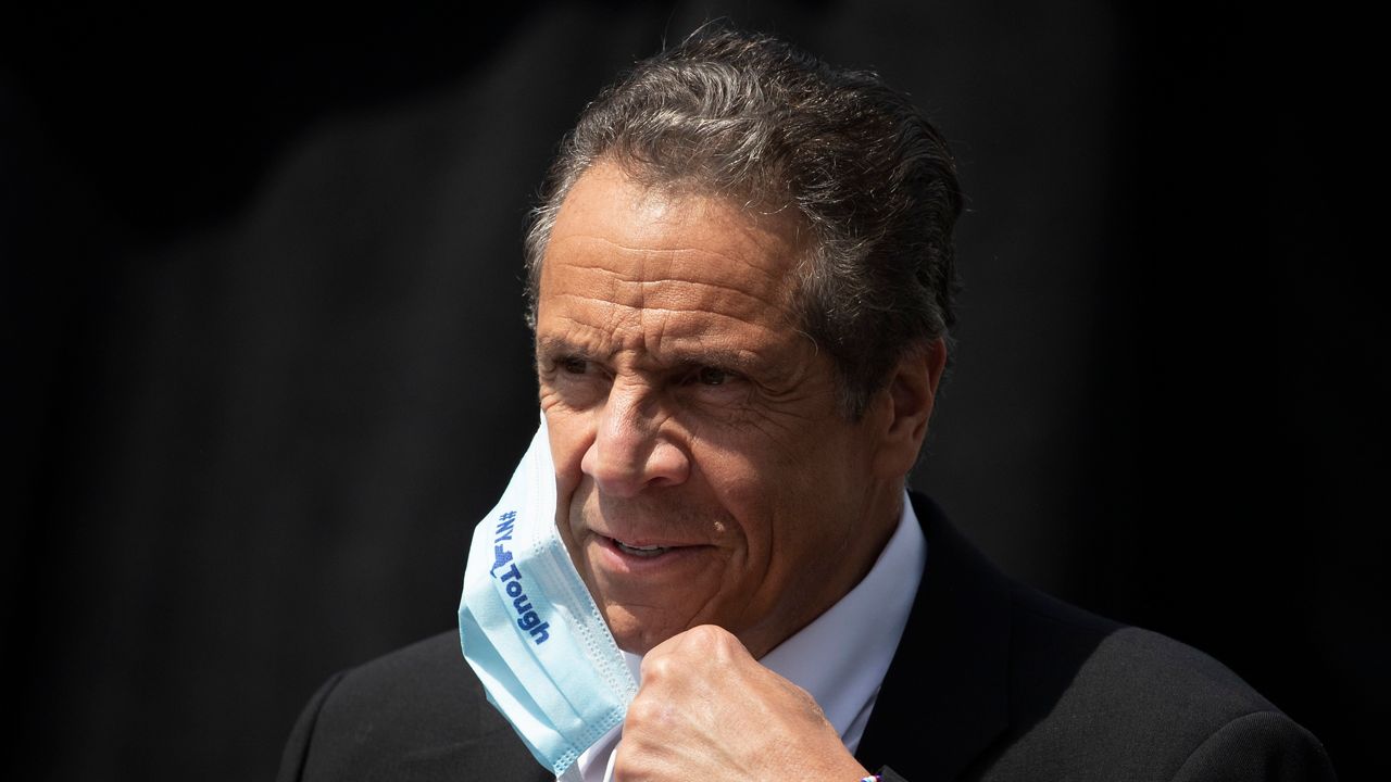 New York Gov. Andrew Cuomo, wearing a black suit jacket, a white dress shirt, and pulling down a sky blue medical mask