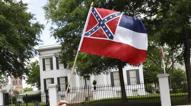 In this April 25, 2020 photograph, a small Mississippi state flag is held by a participant during a drive-by "re-open Mississippi" protest past the Governor's Mansion, in the background, in Jackson, Miss. This current flag has in the canton portion of the banner the design of the Civil War-era Confederate battle flag, that has been the center of a long-simmering debate about its removal or replacement. (AP Photo/Rogelio V. Solis)