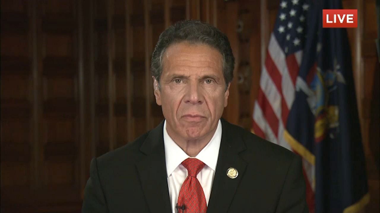 New York Gov. Andrew Cuomo, wearing a black suit jacket, a white dress shirt, and a red tie, sits in front of American and New York state flags.