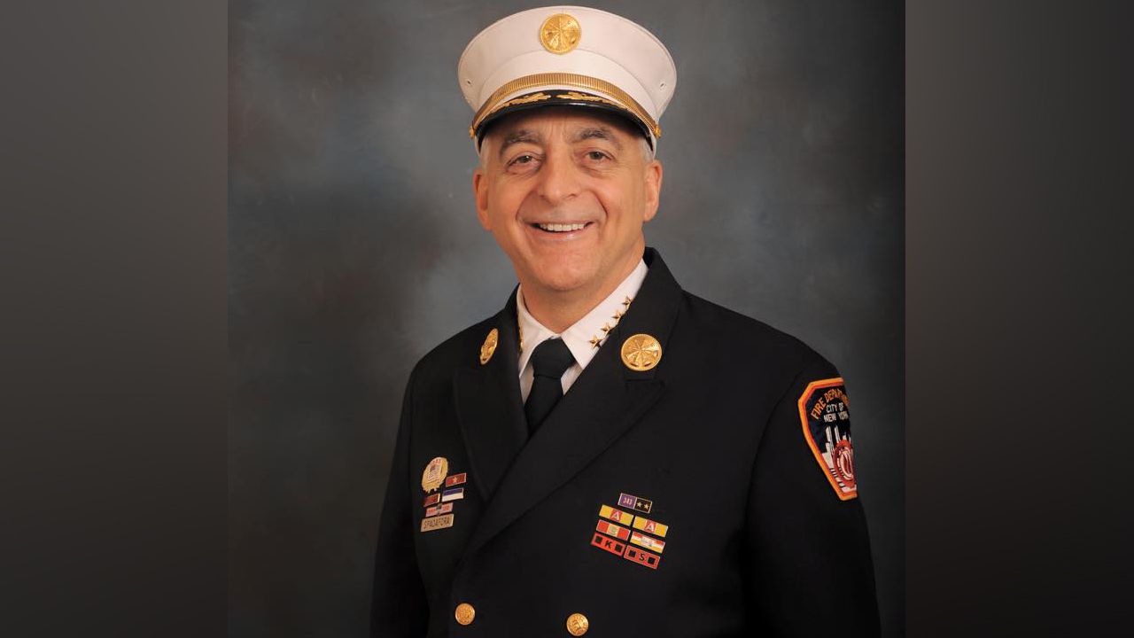 A man wears a black FDNY uniform and a white FDNY hat. Numerous badges and pins are attached to his uniform.