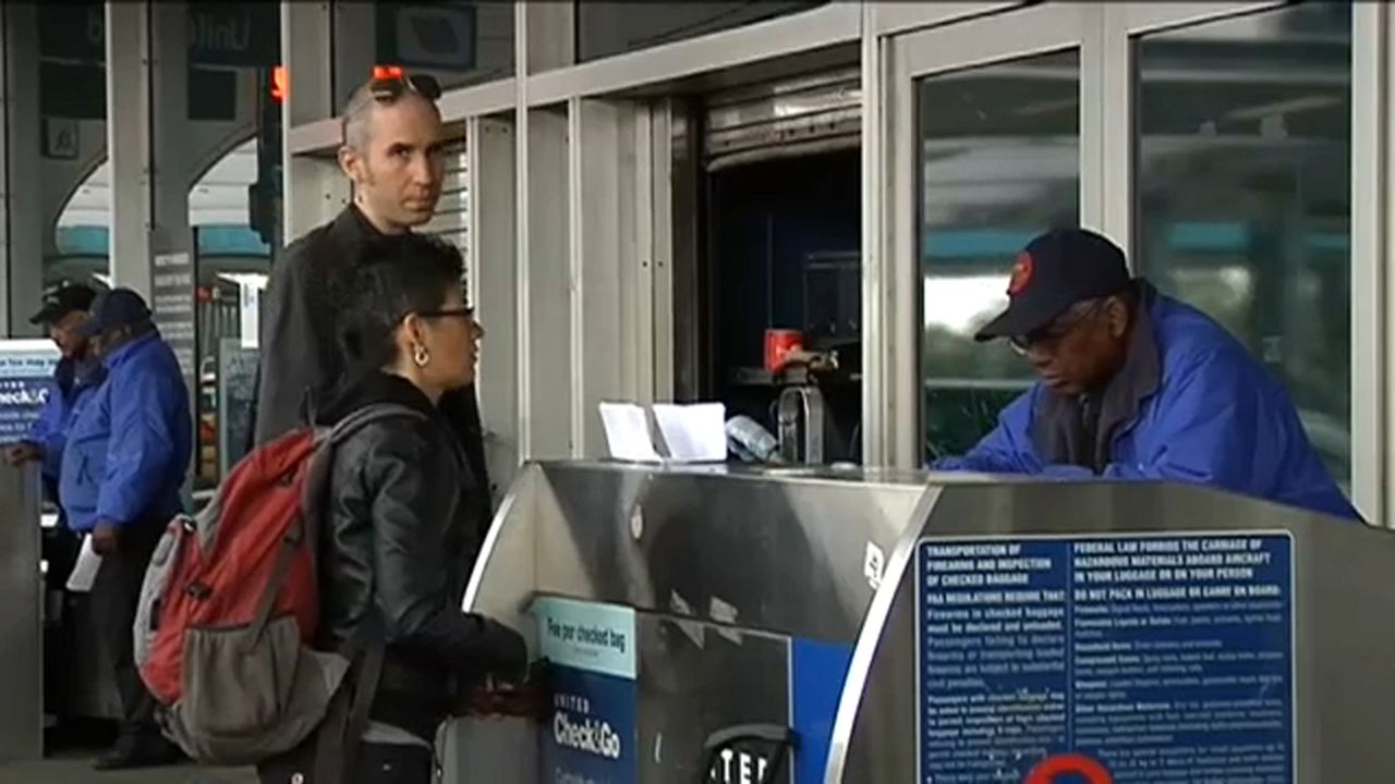 A person, right, wearing a black cap and a blue jacket, stands in front of a glass gate and in front of a metal kiosk. People wearing jackets stand in front of the kiosk.